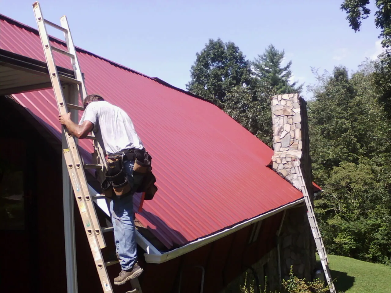 Roofer on a ladder beside a red metal roof and stone chimney — Soffit & Fascia Repair by Roofing Brothers in Franktown